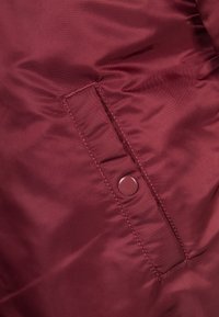 Detailní záběr na kapsu vínově červené látkové bundy s obdélníkovým klopou, která je zajištěná kulatým patentním knoflíkem.