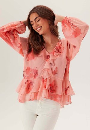 Femme souriante aux longs cheveux bruns portant un chemisier rose à volants fleuri avec des manches bouffantes transparentes et un pantalon blanc, posant avec les bras levés.