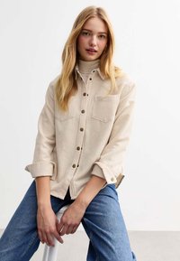Beige corduroy shirt with two chest pockets, button-up front, and rolled sleeves. Worn over a light turtleneck and paired with blue jeans.