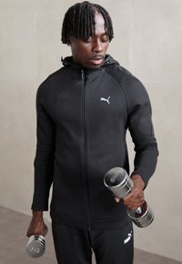 Giacca sportiva nera con zip e cappuccio, caratterizzata da un logo bianco. Una persona tiene dei pesi silver, in piedi contro uno sfondo neutro.