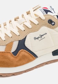 Beige sneaker with suede and textile upper, navy and tan accents, cream laces, and a textured rubber sole. Logo on the side.