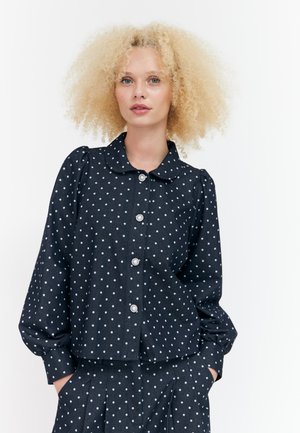 Femme aux cheveux blonds bouclés portant un chemisier noir à manches longues et un pantalon à pois blancs avec boutons en perle, mains dans les poches.