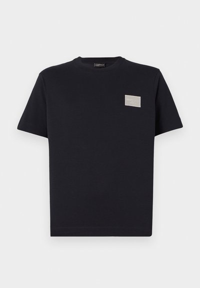 Black cotton t-shirt with short sleeves and a crew neckline. Features a rectangular silver logo patch on the left chest.
