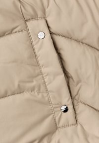 Beige polstret jakke i stof med diamantvattering, med sølvtrykknap-detalje. Glat tekstur med synlige søm-linjer.
