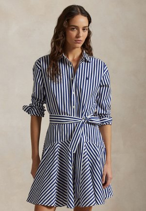 Žena oblečená v modro-bielych vertikálnych pásikavých šatách s golierom, so zdvihnutými dlhými rukávmi a opaskom previazaným v páse.