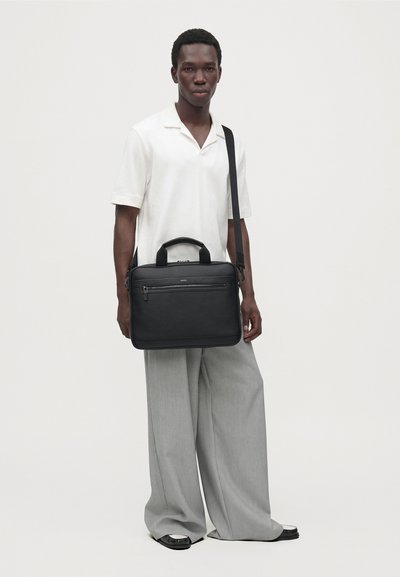 Un homme en polo blanc et en pantalon gris ample présente un sac porté à l’épaule noir, doté d’une poignée supérieure et d’une poche avant zippée.