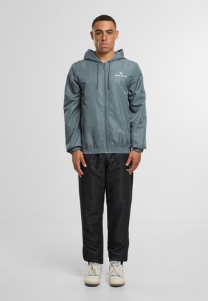 Vaalean teal huppari, jossa on sileä pinta, vetoketjulukko ja joustava helma, yhdistettynä mustiin housuihin ja valkoisiin kenkiin. Minimimerkintä näkyvissä.