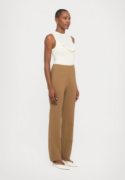 Cowl-neck cream top paired with high-waisted, wide-leg brown trousers and white heels, showcasing smooth fabric and tailored fit.
