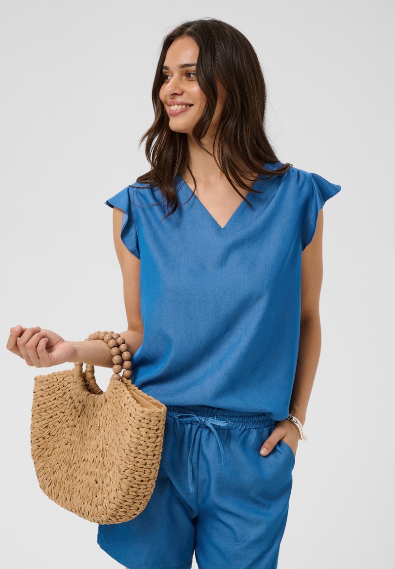 Femme en haut bleu sans manches et short tenant un sac à main beige tissé avec des poignées rondes en bois, souriante et regardant sur le côté.