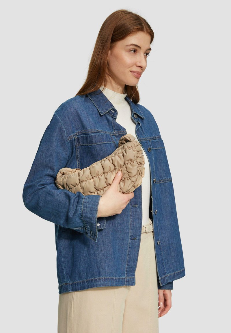 Beige, gesteppte Clutch-Tasche, die in einer Hand gehalten wird, mit einem abgerundeten Design. Kombiniert mit einer Jeansjacke und einem hellen Oberteil, mit sichtbaren beigen Hosen.