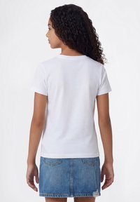 Joven con cabello rizado que lleva una camiseta blanca de manga corta lisa y una falda vaquera azul, de espaldas a la cámara.