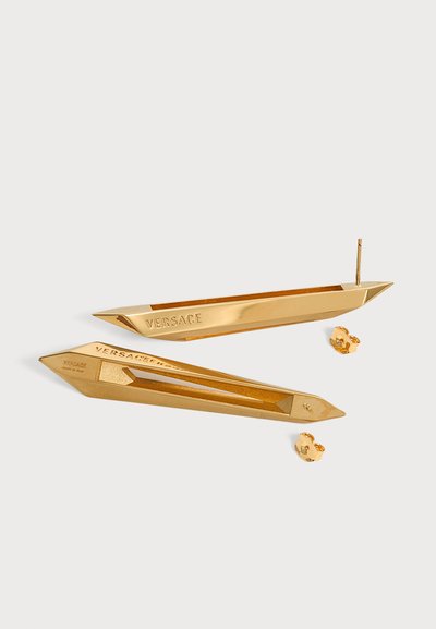Paire de boucles d'oreilles Versace ton or, en forme de barre géométrique allongée avec des extrémités pointues et des attaches papillon sur une surface blanche.