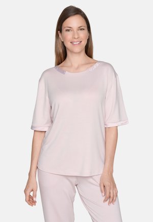 Lächelnde Frau mit langen braunen Haaren, die ein hellrosa Kurzarmshirt und passende Hosen vor einem schlichten weißen Hintergrund trägt.