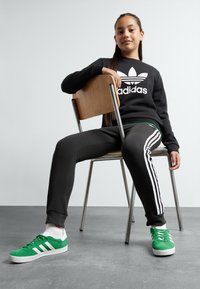 Juodas džemperis su baltu Adidas logotipu, juodos sportinės kelnės su baltais dryželiais ir žali suede sportbačiai su baltais akcentais, sėdinys ant metalinės kėdės.