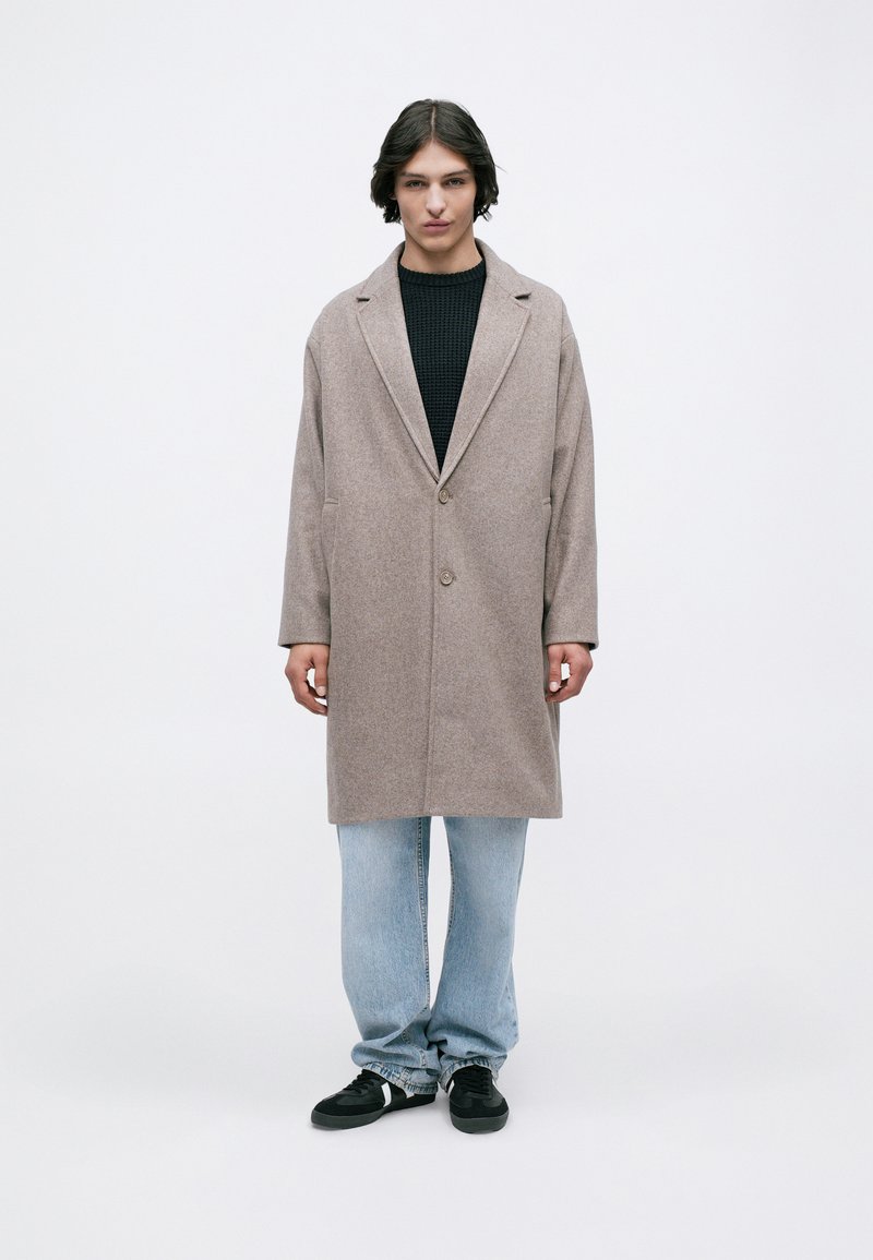 Light brown wool coat with a notched collar, single button closure, and side pockets, worn over a textured black sweater and light jeans.