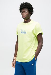 Neónovo žlté bavlnené tričko s vyhrnutými rukávmi, na prednej strane s modrým vyšívaným logom "Café Swoosh". Nosí sa s modrými nohavicami.