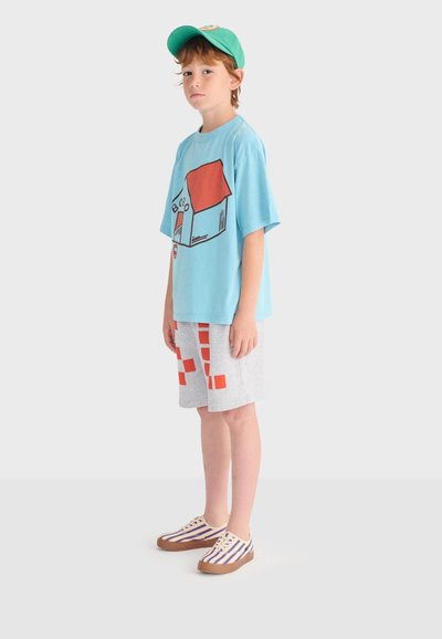 Enfant portant une casquette verte, un T-shirt bleu oversize avec un motif de maison, un short gris à carreaux rouges, et des baskets rayées, debout de profil.