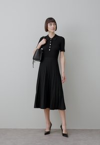 Kvinna med kort mörkt hår som bär en svart, kortärmad, veckad klänning, svarta klackskor och håller en svart handväska på axeln.