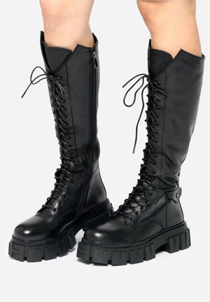 Černé vysoké kozačky na šněrování po kolena s robustními podrážkami a bočními zipy, nošené na holených nohou.