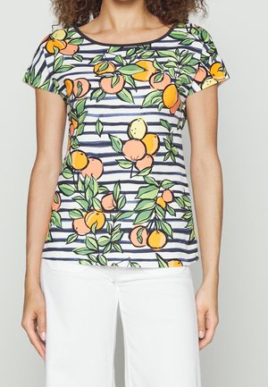 Femme portant une chemise à manches courtes rayée bleu marine et blanc avec un motif de fruits feuillus orange, pêche et vert, associée à un pantalon blanc.