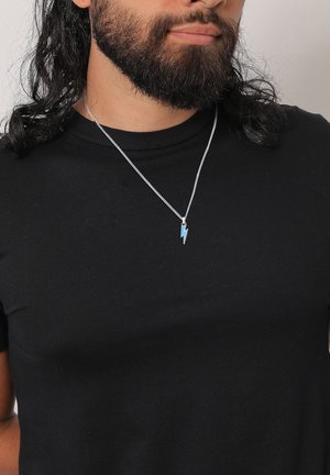 Silver chain necklace featuring a small blue lightning bolt pendant, worn over a black t-shirt. Textured hair and beard visible.