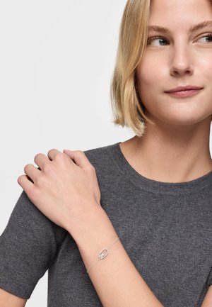 Młoda kobieta z krótkimi blond włosami, ubrana w szary, dziany top i delikatną srebrną bransoletkę ze wzorem owalnym i krzyżykiem na nadgarstku.