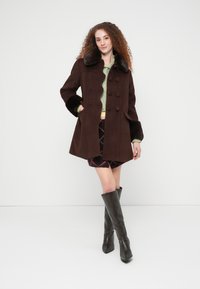 Καφέ μάλλινο παλτό με τεχνητό γούνινο γιακά και μανσέτες, διπλό στήθος, σε συνδυασμό με καρό φούστα και δερμάτινες μπότες έως το γόνατο.