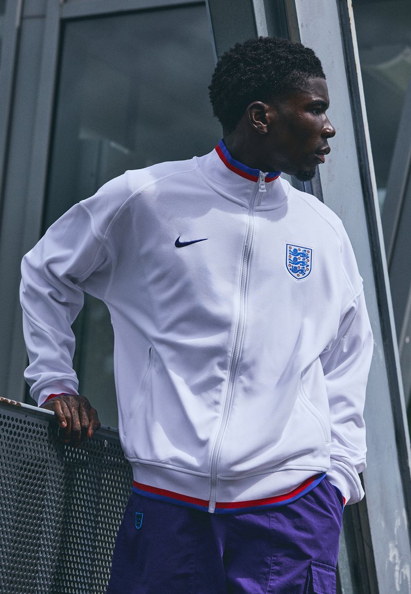 Hvit jakke med glidelås og høy krage, rød og blå kant, Nike-logo på venstre bryst, Englands emblem på høyre. Brukt sammen med lilla shorts.