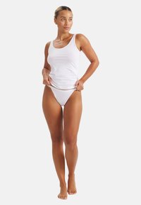 Žena stojící naboso, oblečená v bílém tílku a odpovídajícím bílém spodním prádle, upravující lem svého topu, na jednobarevném světlém pozadí.