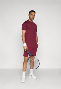 Burdeos spalvos sportinis marškinėliai ir šortai, derinami su baltais riešų raiščiais ir sportbatiais. Laiko teniso raketę su mėlynu akcentu.