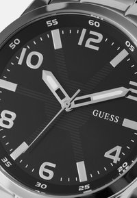 Rundes schwarzes Zifferblatt mit silbernen Stundenmarkierungen und -zeigern. Verfügt über einen strukturierten Hintergrund und das 'GUESS'-Logo. Silbernes Metallband.