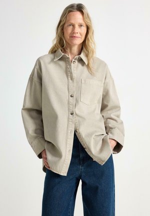 Donna con lunghi capelli biondi che indossa una camicia beige oversize con bottoni e jeans blu scuro con le mani nelle tasche su uno sfondo semplice.