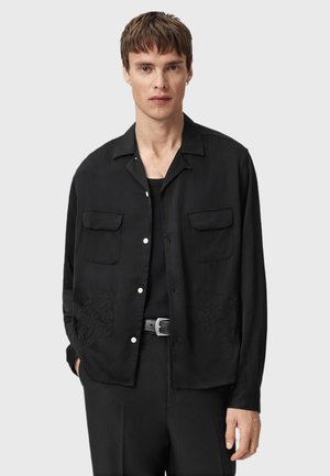 Black shirt made of fabric with a smooth texture, featuring two chest pockets and decorative embroidery at the bottom. Designed with a relaxed fit.