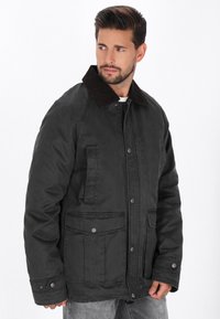 Zwarte gewaxte stofjas met een bruine corduroy kraag, voorzien van twee voorzakken en een knoopsluiting, ontworpen voor warmte en duurzaamheid.