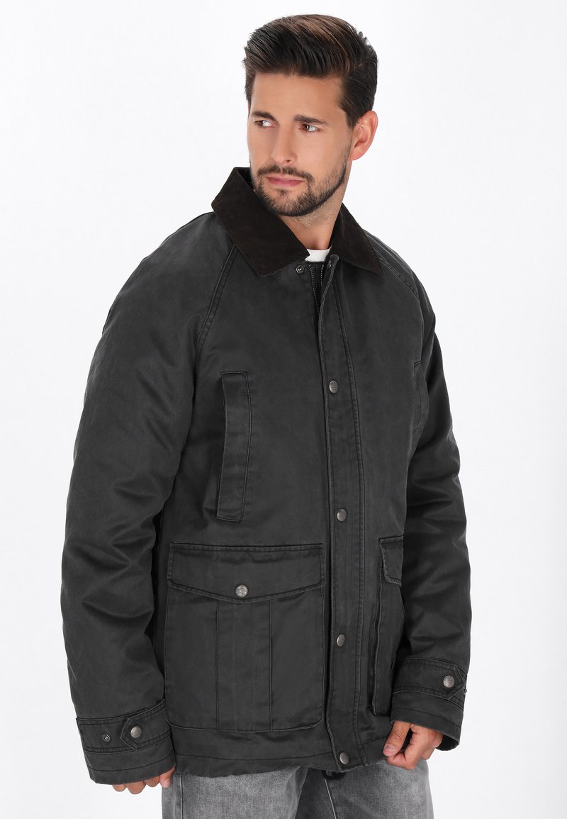 Chaqueta de tela encerada negra con un cuello de pana marrón, que cuenta con dos bolsillos frontales y un cierre de botón, diseñada para proporcionar calidez y durabilidad.