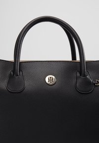 Sac à main en cuir noir avec des poignées arrondies et un petit emblème rond en métal centré entre elles.
