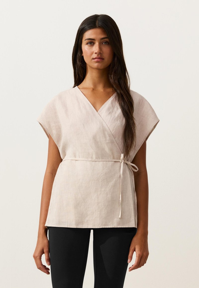 Femme aux longs cheveux foncés portant un haut beige à manches courtes style portefeuille noué à la taille, associé à un pantalon noir, debout devant un fond uni.