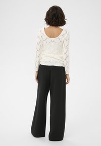 Fehér kötött pulóver gyémánt mintával és mély hátrészettel, magas derekú, fekete bő szárú nadrággal párosítva. Puha tapintás, laza illeszkedés.