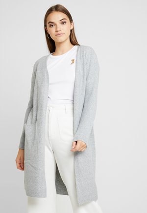 Lichtgrijze gebreide cardigan met open voorkant, lange mouwen en zijzakken. Gecombineerd met witte ribbelbroek en een wit shirt daaronder.