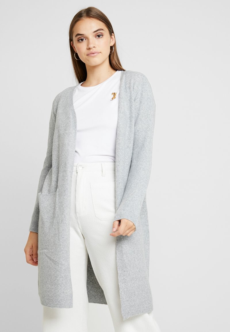 Hellegraue Strickjacke mit offenen Vorderseite, langen Ärmeln und seitlichen Taschen. Kombiniert mit weißen Ripp-Hosen und einem weißen Hemd darunter.