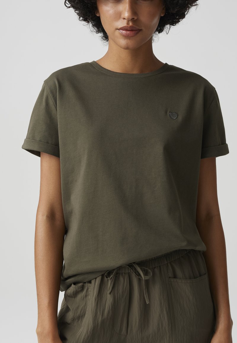 Woman wearing an olive green t-shirt with rolled sleeves and a small heart emblem, paired with matching drawstring pants. Upper body visible.