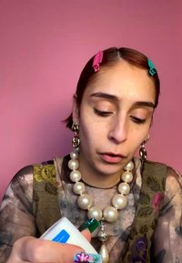 Femme avec des pinces à cheveux roses et vertes applique une lotion d'une bouteille blanche, porte un grand collier de perles, un nail art floral et un haut à motifs devant un mur rose.