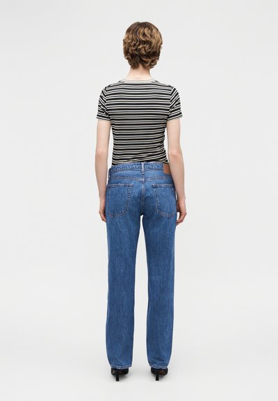 Άτομο με κοντά σγουρά μαλλιά, φοράει ριγέ μπλούζα μαύρου και λευκού χρώματος, μπλε τζιν ίσιας γραμμής και μαύρα παπούτσια με τακούνι, στέκεται με την πλάτη προς το μέρος του θεατή.