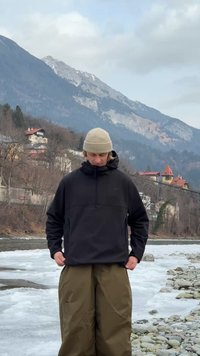 Person iført beige hue og sort jakke står på en stenfyldt flodbred med isende vand og sneklædte bjerge i baggrunden.