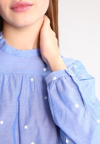 Blouse bleu clair ornée de motifs d'étoiles blanches, tissu doux, détails froncés au niveau de l'encolure et des manches, et col rond.