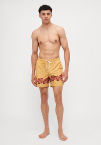Schwimmshorts in hellgelb mit einem gemusterten roten und schwarzen Design am Saum. Sie verfügen über einen elastischen Bund mit Kordelzug.