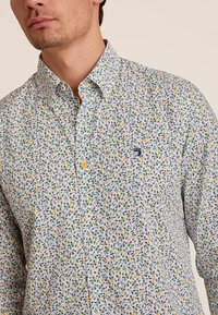 Witte button-up shirt van stof met een veelkleurig bloemenpatroon. Heeft een spitse kraag en een klein logo op de borst.