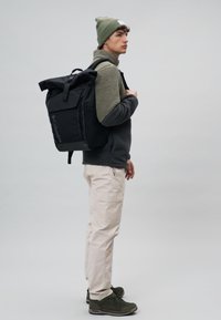 Black backpack with multiple compartments, adjustable straps, and embroidered logo; worn with a green fleece and beanie, light pants, and green shoes.