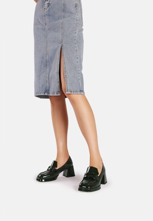 Mocassini neri in pelle verniciata con tacco spesso, dotati di una fascia decorativa e punta arrotondata, abbinati a una gonna a tubino in denim.