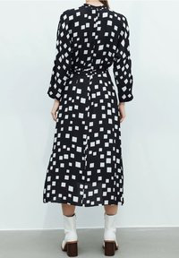 Schwarzes Kleid mit langen Ärmeln und taillierter Taille, mit einem weißen quadratischen Muster. Glattes Material, knöchellang, ohne sichtbare Beschläge.